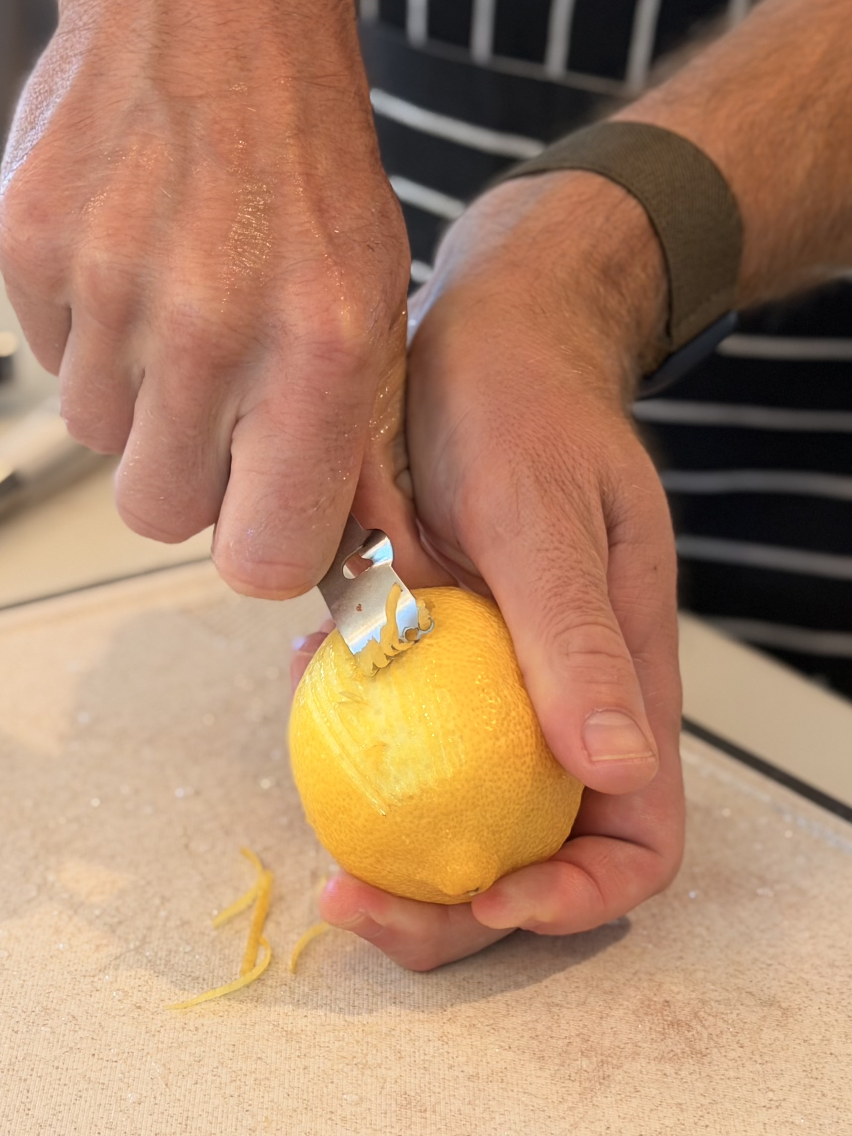 A person's hands use a zester on a whole lemon over a wooden cutting board, with curls of yellow zest falling onto the board in a