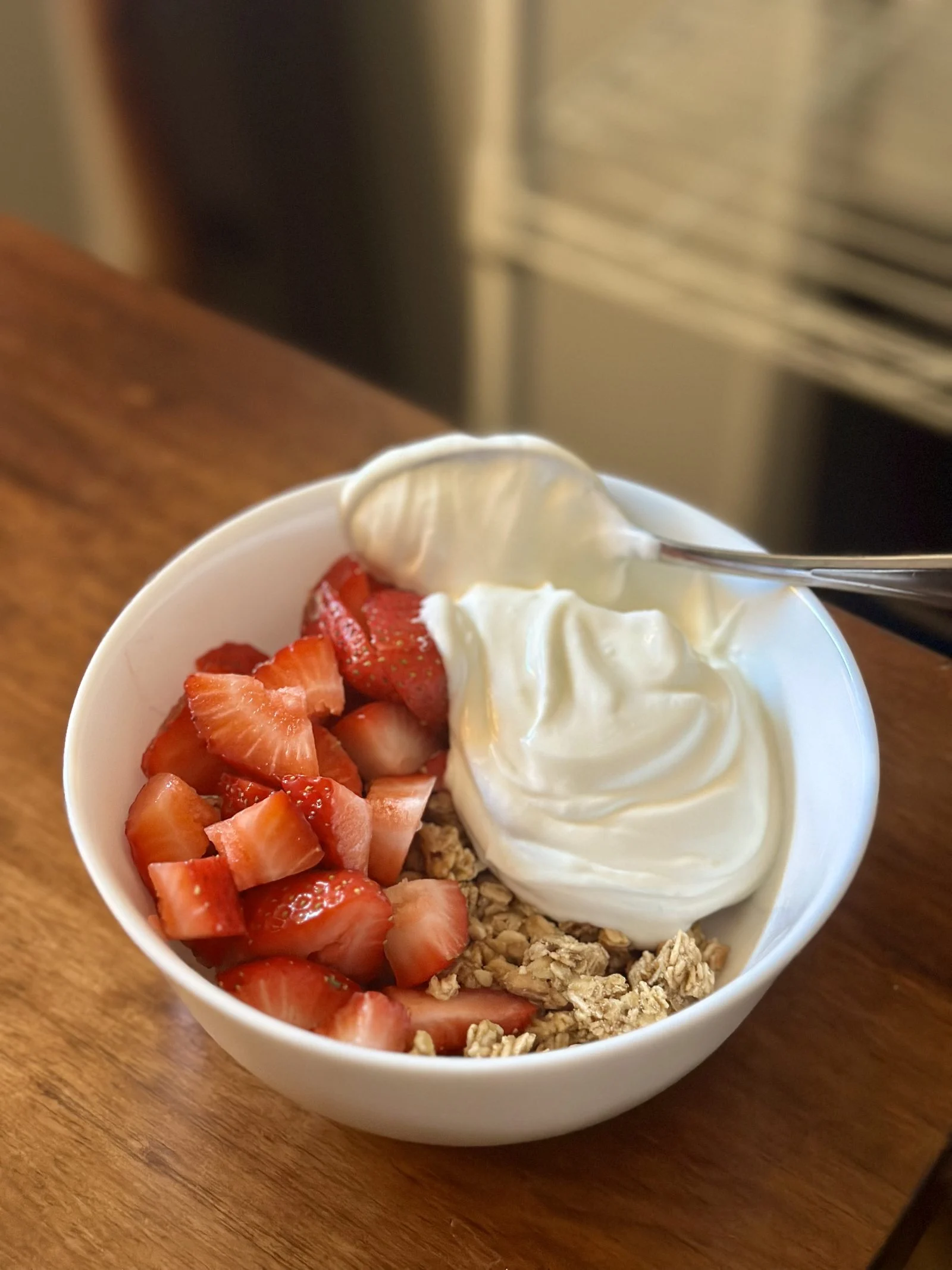 A white bowl holding a scoop of thick yogurt with chopped strawberries and granola, a spoon resting in it, on a wooden table.