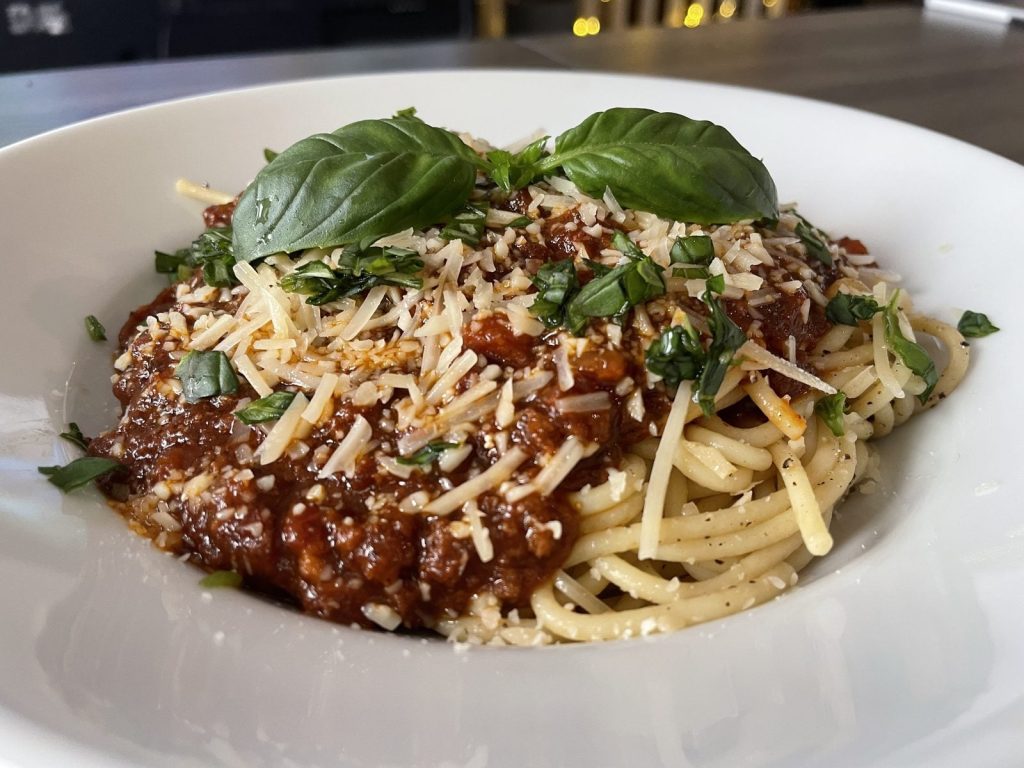 A white bowl of spaghetti topped with a chunky meat ragu, shredded parmesan and fresh basil leaves.