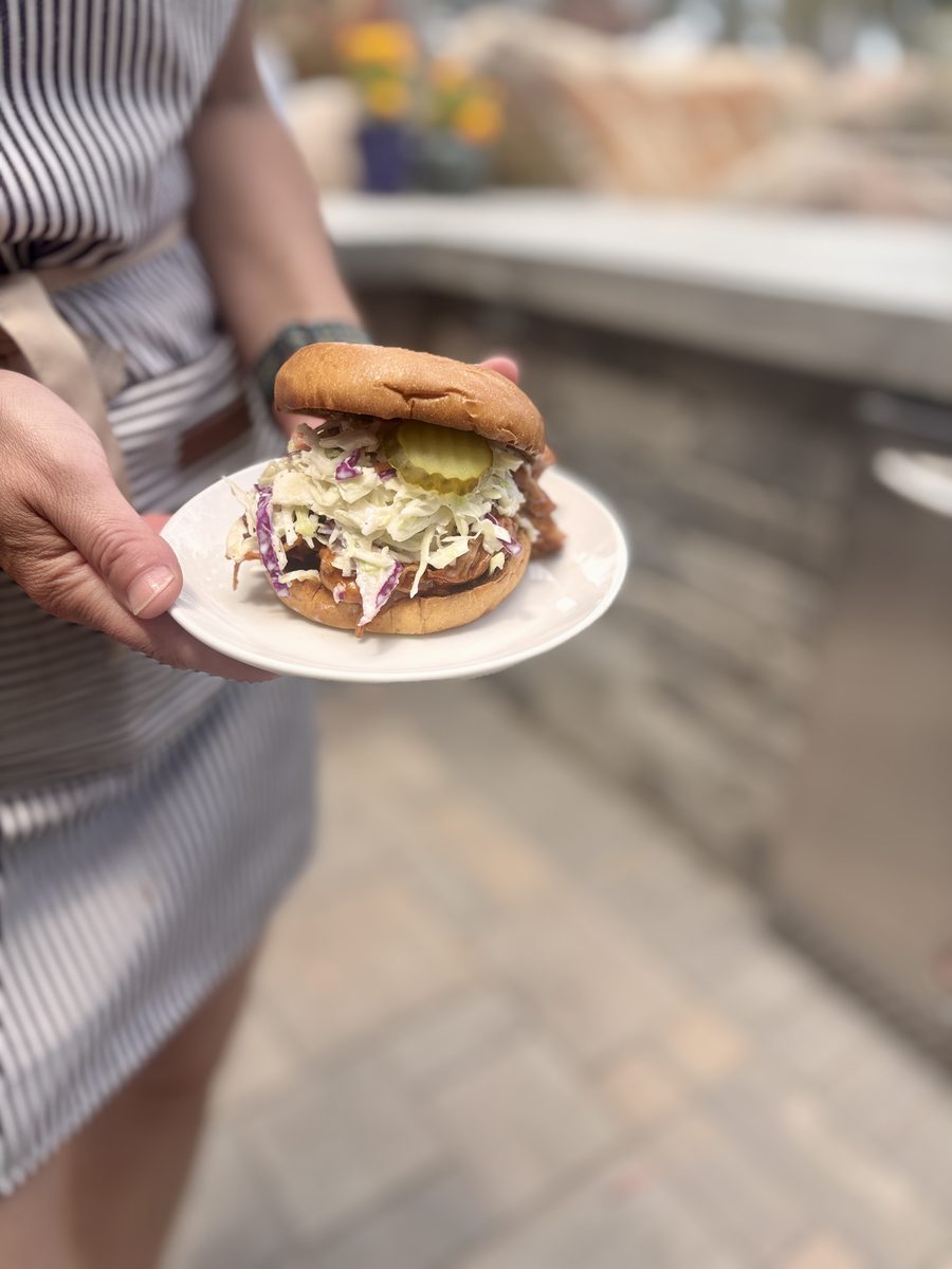 A woman in a striped apron holds a small white plate with an assembled BBQ pulled-chicken sandwich topped with slaw and pickle sli