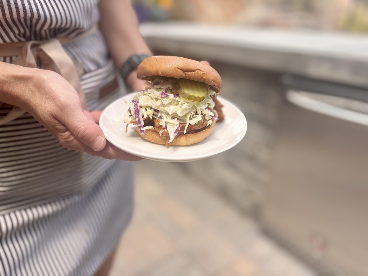 Hands hold out a white plate with a tall BBQ pulled-chicken sandwich stacked with slaw and pickles, outdoors in bright daylight.