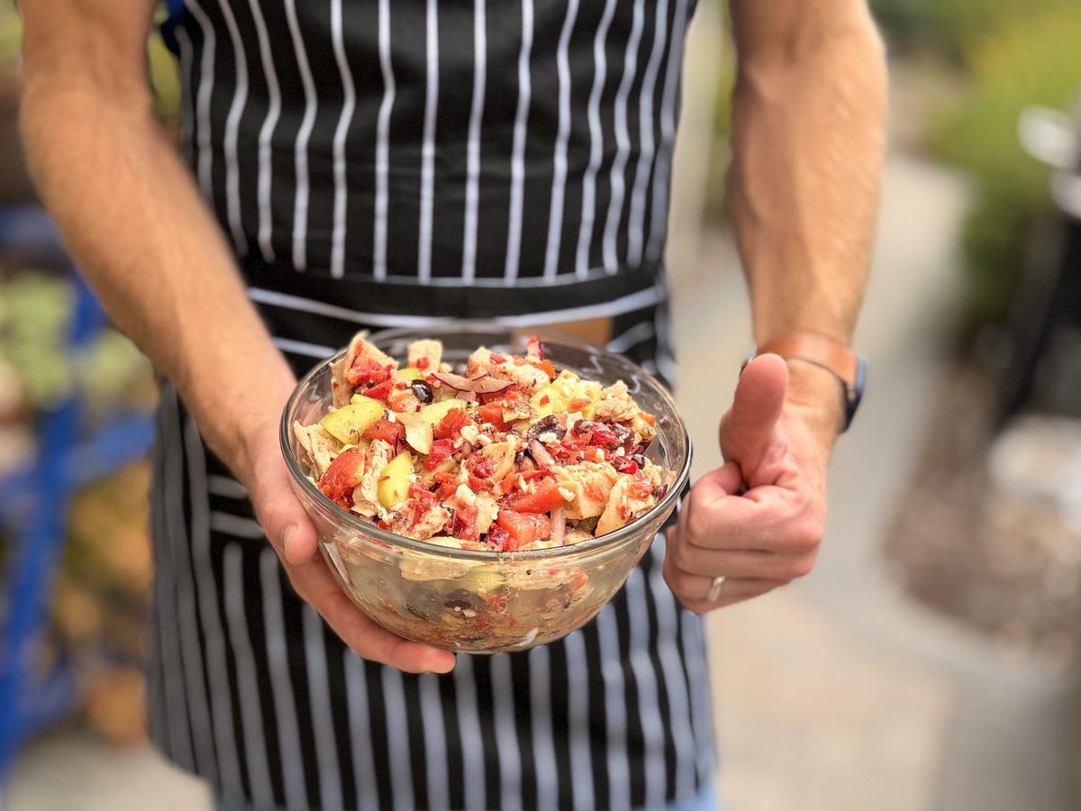A man in a striped apron holds a large glass bowl of chopped Greek-style salad with tomato, cucumber, feta and pieces of grilled c