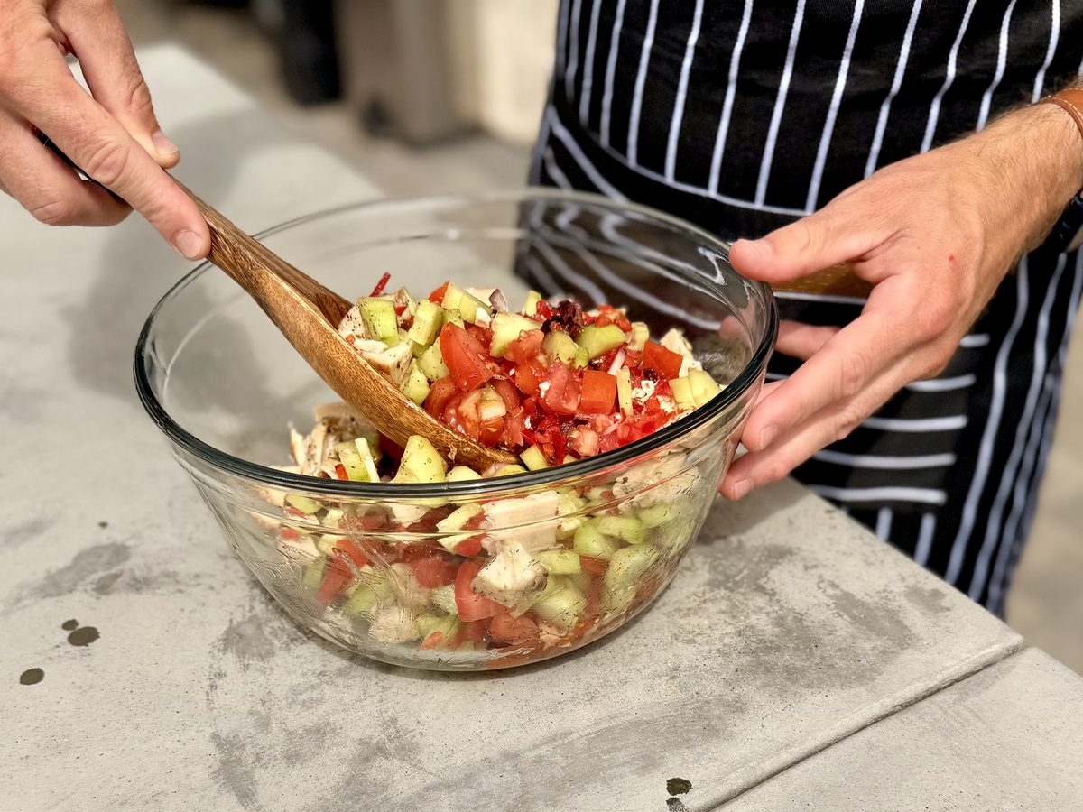 Hands toss a chopped salad of tomato, cucumber and pale chicken chunks with a wooden spoon in a glass mixing bowl on a concrete co