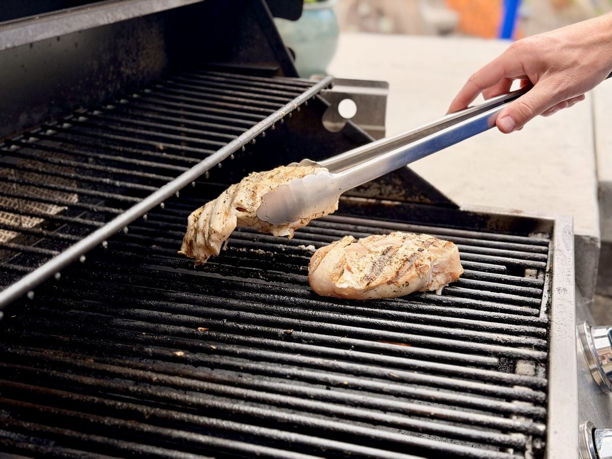 A hand with tongs turns two chicken breasts on the grates of an open gas grill outdoors.