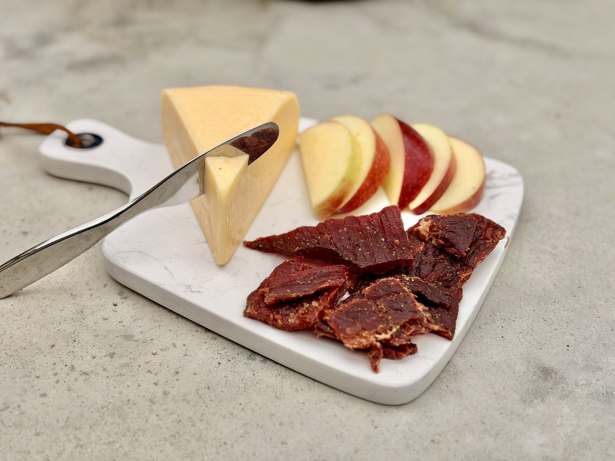 A white marble serving board on a concrete surface holding a wedge of gouda with a cheese knife, fanned red apple slices and a pil