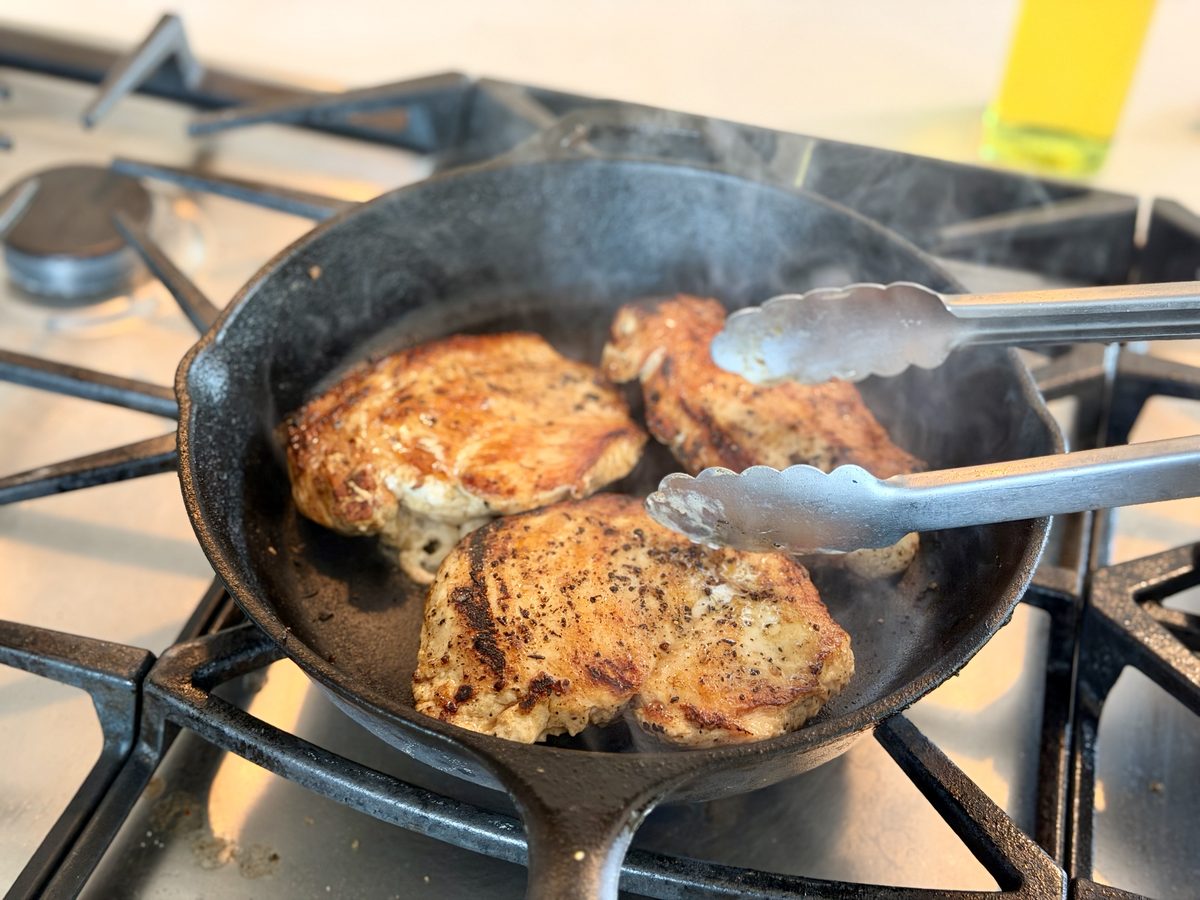 Seasoned chicken breasts browning in a cast iron skillet on a gas range, with tongs resting in the pan.