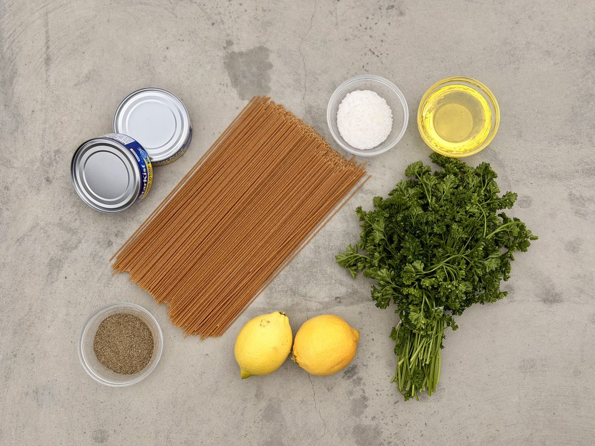 Overhead ingredient flat lay on grey concrete: two sealed tin cans, dry whole wheat spaghetti, a bunch of parsley, two lemons and