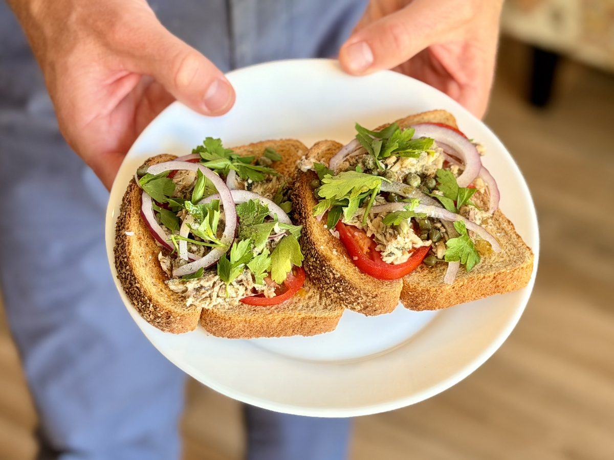 Two hands hold a white plate with two open-faced whole-grain toasts topped with mashed sardines, tomato slices, red onion rings, c