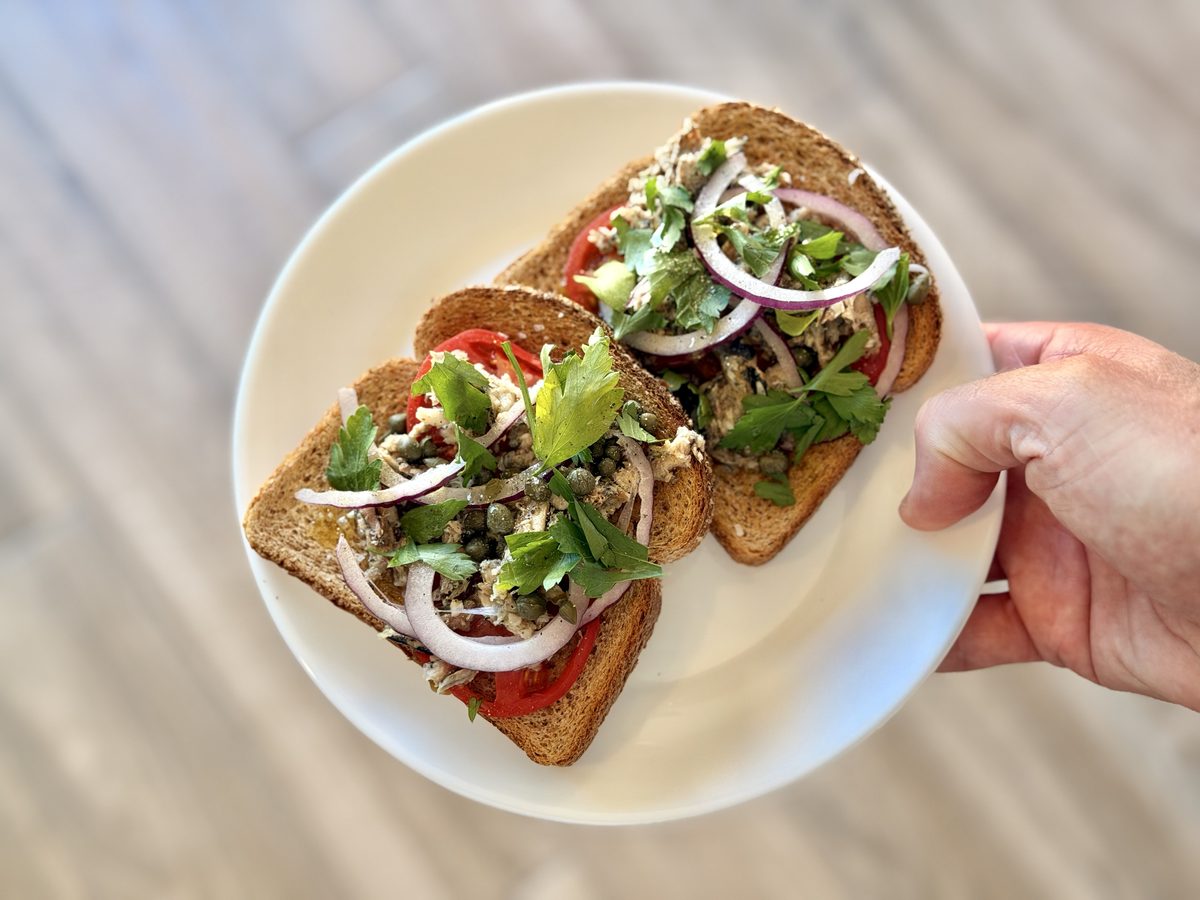A hand holds a white plate of two open-faced sardine toasts with tomato, red onion, capers and parsley, shot from above over a woo