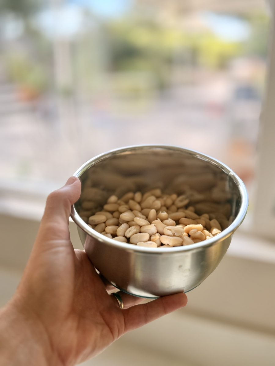 A hand holds a small stainless bowl of drained white beans up toward the camera, with a blurred outdoor kitchen behind.