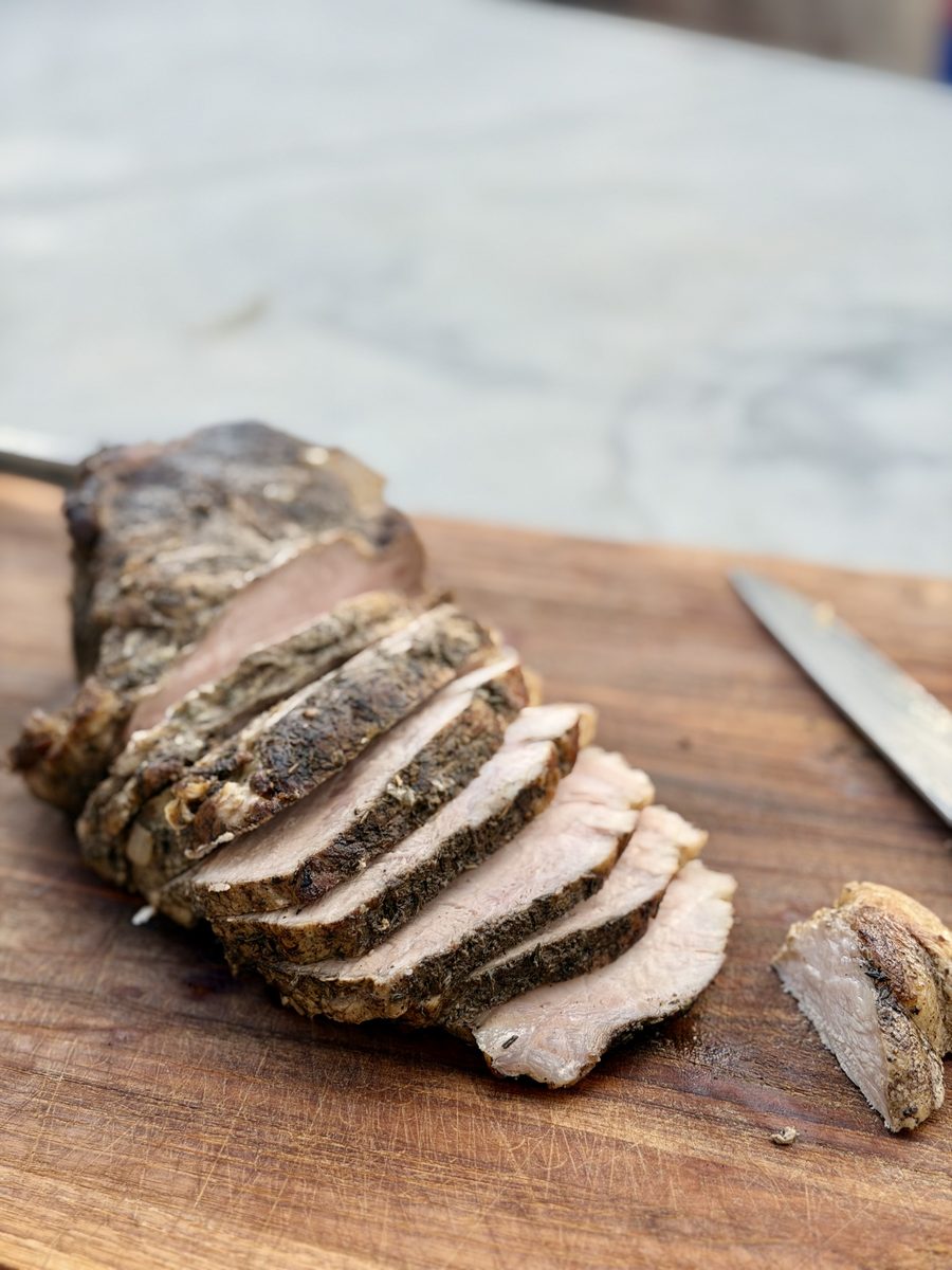 A herb-crusted roast of pork loin, sliced into thick medallions, resting on a wooden cutting board with a carving knife beside it 