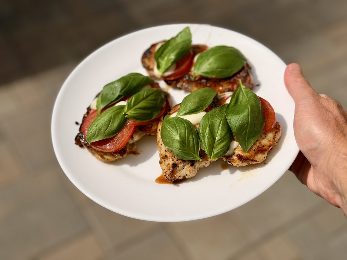 A hand holds a white oval platter of two chicken breasts topped with tomato slices, melted mozzarella and whole basil leaves over 