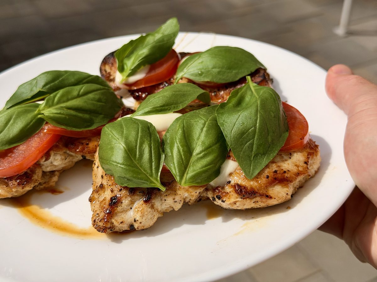 Close overhead of a held white platter of caprese-style chicken breasts with tomato, melted mozzarella and basil leaves.