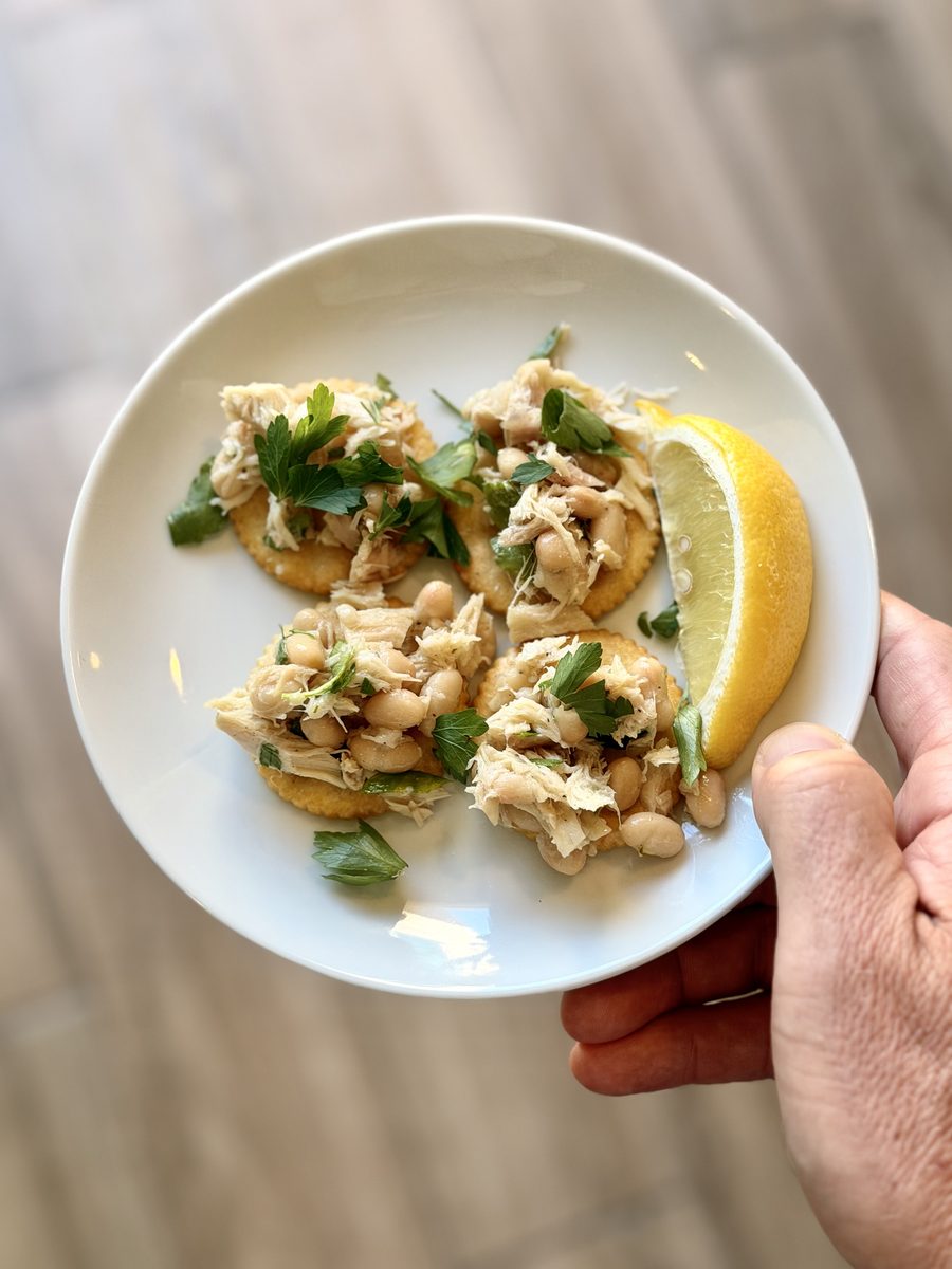 A hand holds a white plate of round crackers topped with flaked tuna, white beans and parsley with a lemon wedge, shot from above