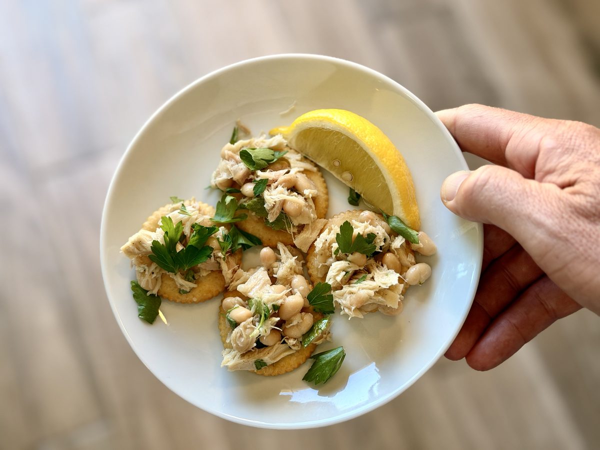 Close overhead of a held white plate of crackers piled with flaked tuna, cannellini beans and fresh parsley next to a lemon wedge.