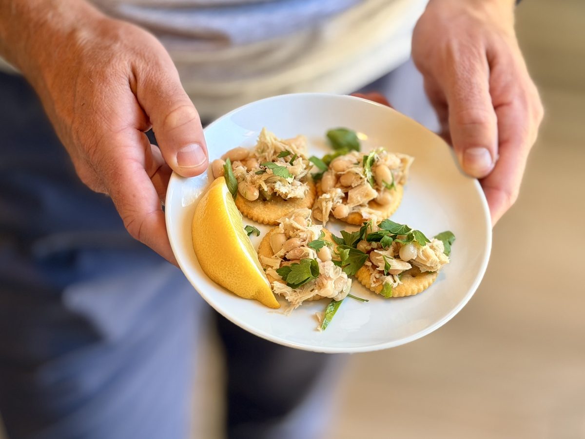 A man in jeans holds a white plate of tuna and white bean topped crackers with a lemon wedge at waist height.