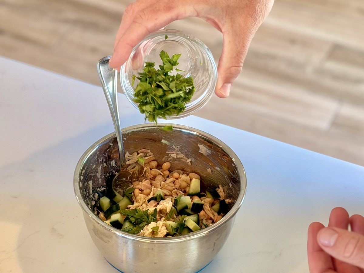 Hands tip chopped parsley from a glass bowl into a stainless mixing bowl of flaked tuna, beans and diced cucumber on a white count