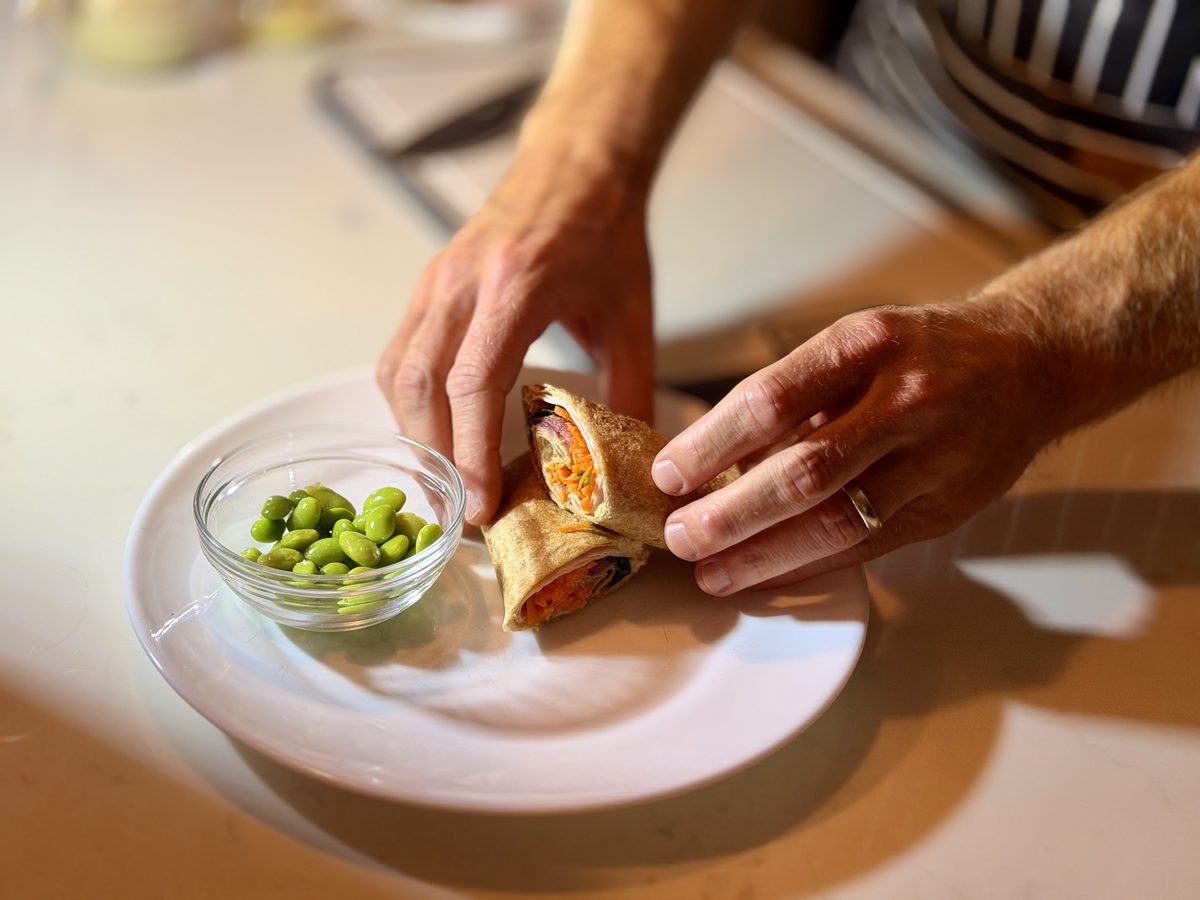 Hands setting two halves of a finished rolled wrap onto a white plate next to a small glass bowl of edamame on a kitchen counter.