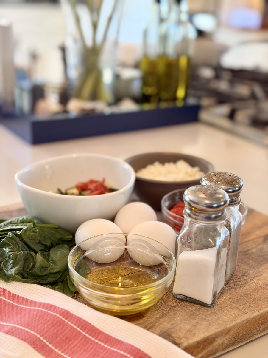 Ingredients arranged on a wooden board on a counter: baby spinach, three brown eggs, diced tomato, crumbled feta, olive oil and a 