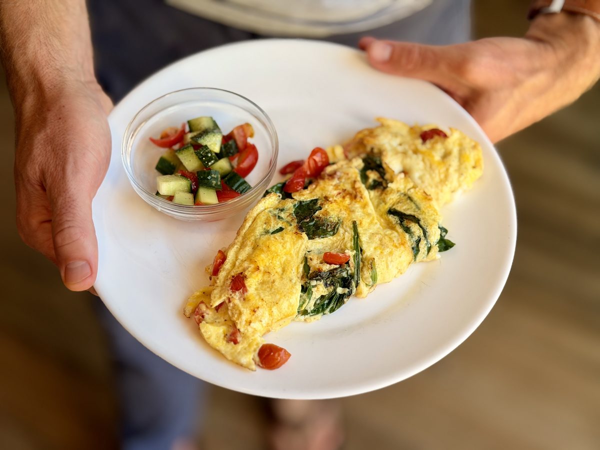 A person holds a white plate with a folded spinach and tomato omelet beside a small glass bowl of chopped cucumber-tomato salad.
