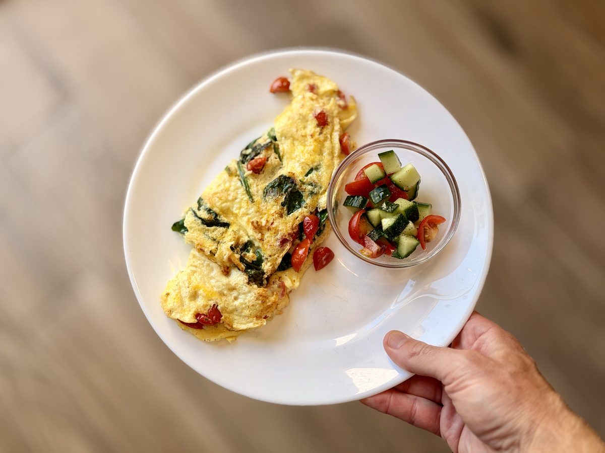 Overhead of a held white plate with a folded veggie omelet and a small glass bowl of cucumber, tomato and feta salad, over a wood