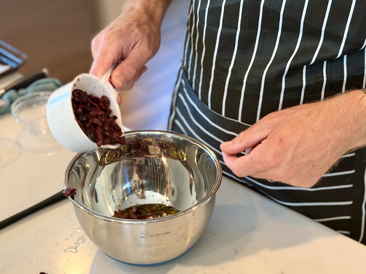 A cook in a striped apron tips chopped dark olives from a white bowl into a large stainless mixing bowl on a kitchen counter.