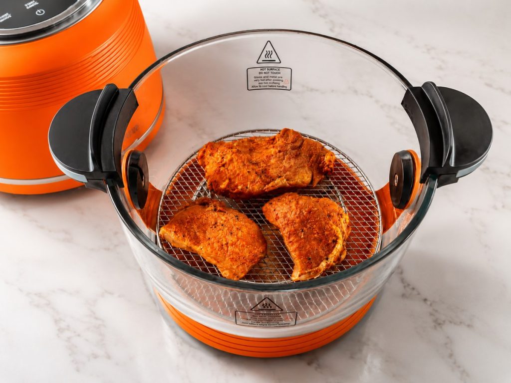 Three pieces of red-spice-rubbed cooked chicken sit on the mesh rack inside the glass bowl of an orange countertop air fryer on a marble surface.
