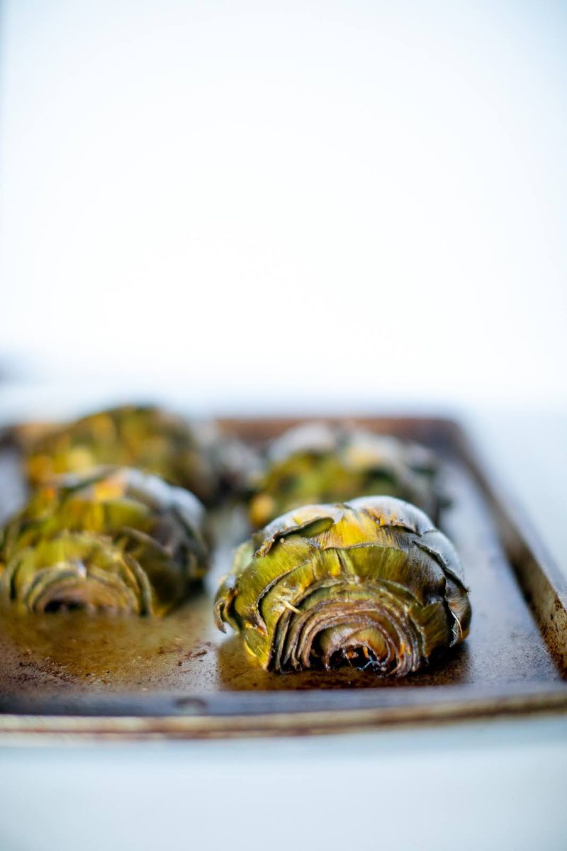 Three roasted artichoke halves cut side up on a metal sheet pan on a white counter, bright high-key background.