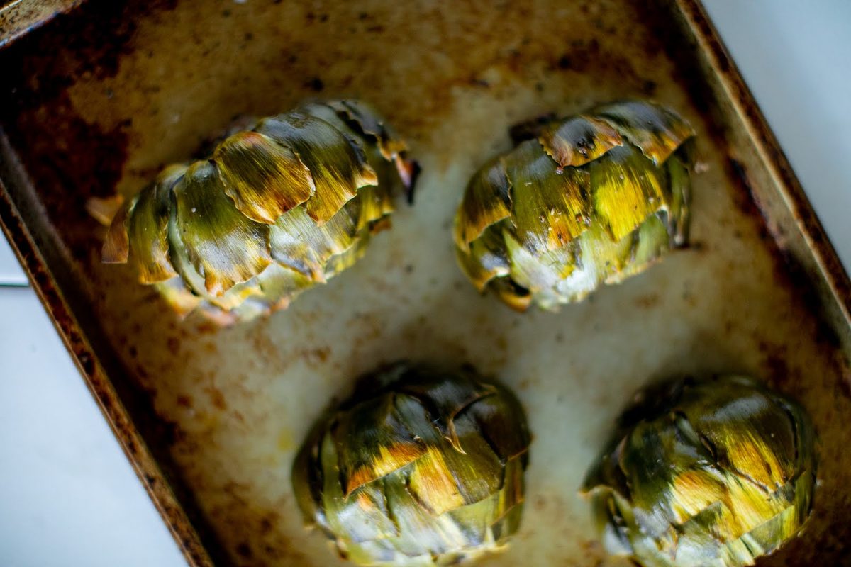 Four halved artichokes browned and bubbling in a creamy cheese sauce in a dark roasting dish straight from the oven.