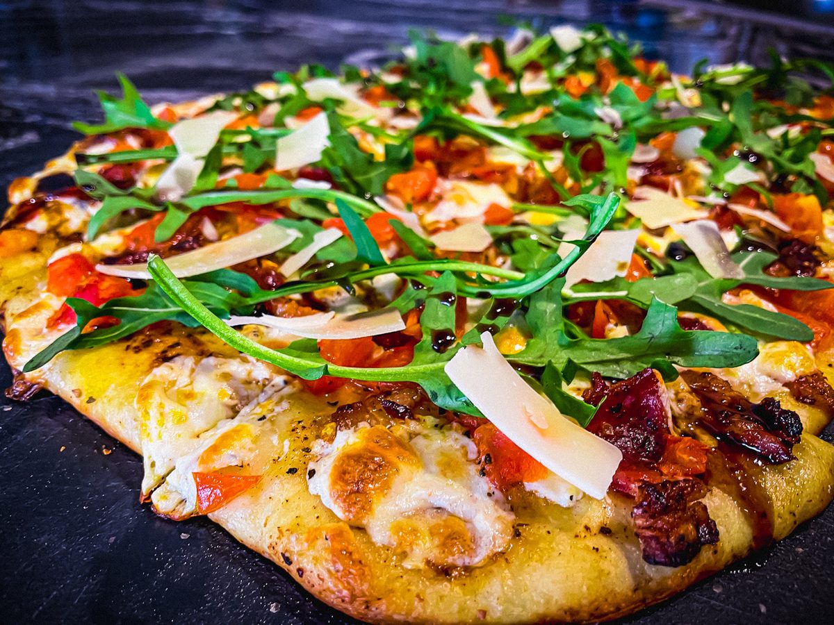 Close-up of a rectangular flatbread pizza with melted cheese, diced tomato, shaved parmesan, fresh arugula and crisp dark red curl