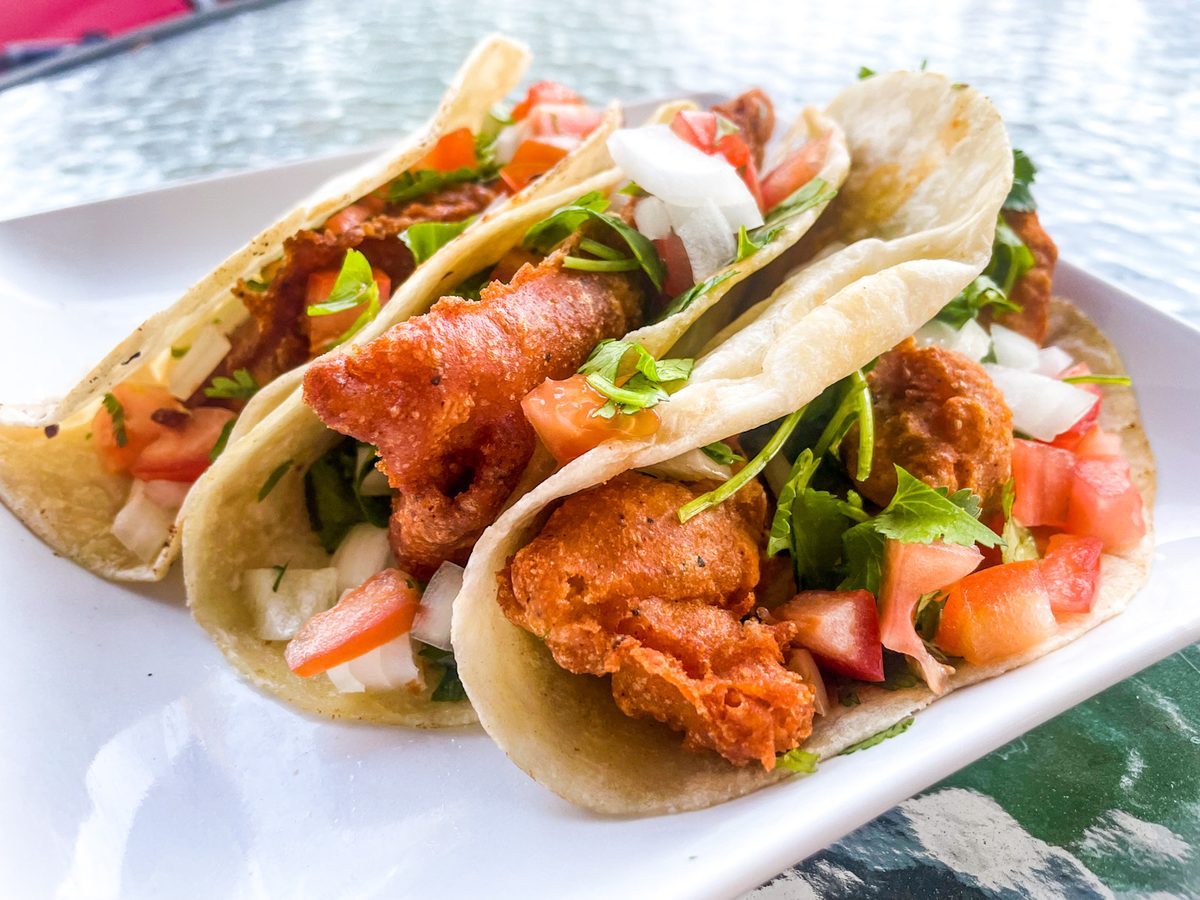 Three flour tortilla tacos filled with battered fried fish, shredded cabbage, pico de gallo and white crema, lined up on a white r