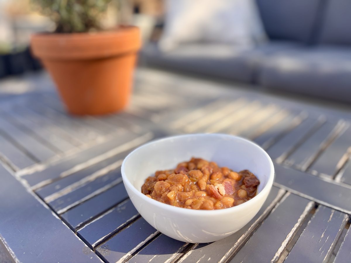 A small white bowl of brown beans in sauce sitting on a slatted grey outdoor table with a terracotta pot behind.