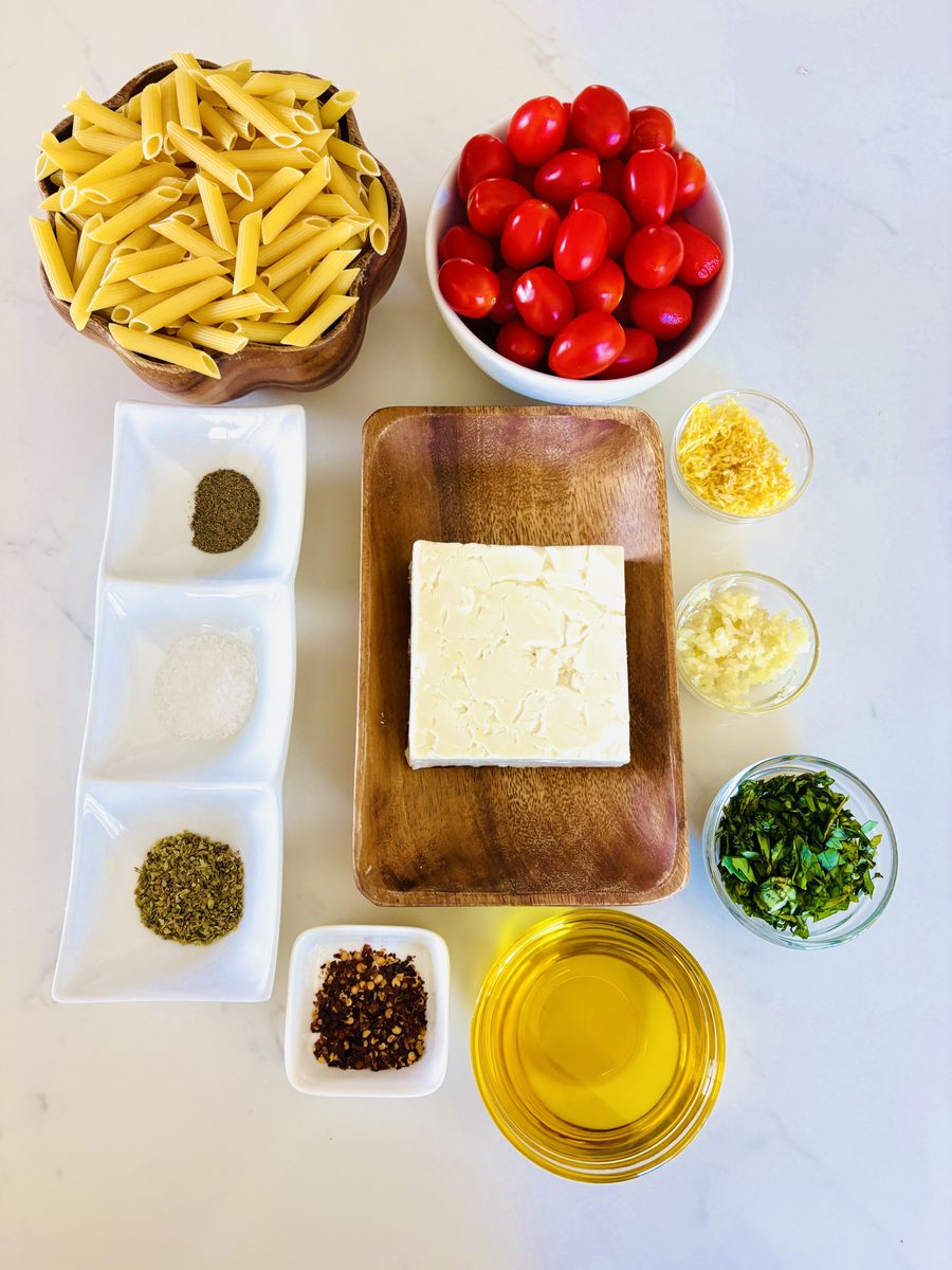 Overhead ingredient flat lay on white marble: a wooden bowl of dry penne, a bowl of cherry tomatoes, a block of feta on a wooden b
