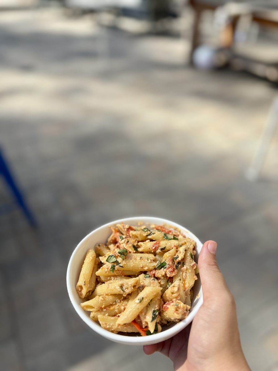 Hands hold a small white bowl of penne in creamy feta-tomato sauce, outdoors with a blurred patio behind.