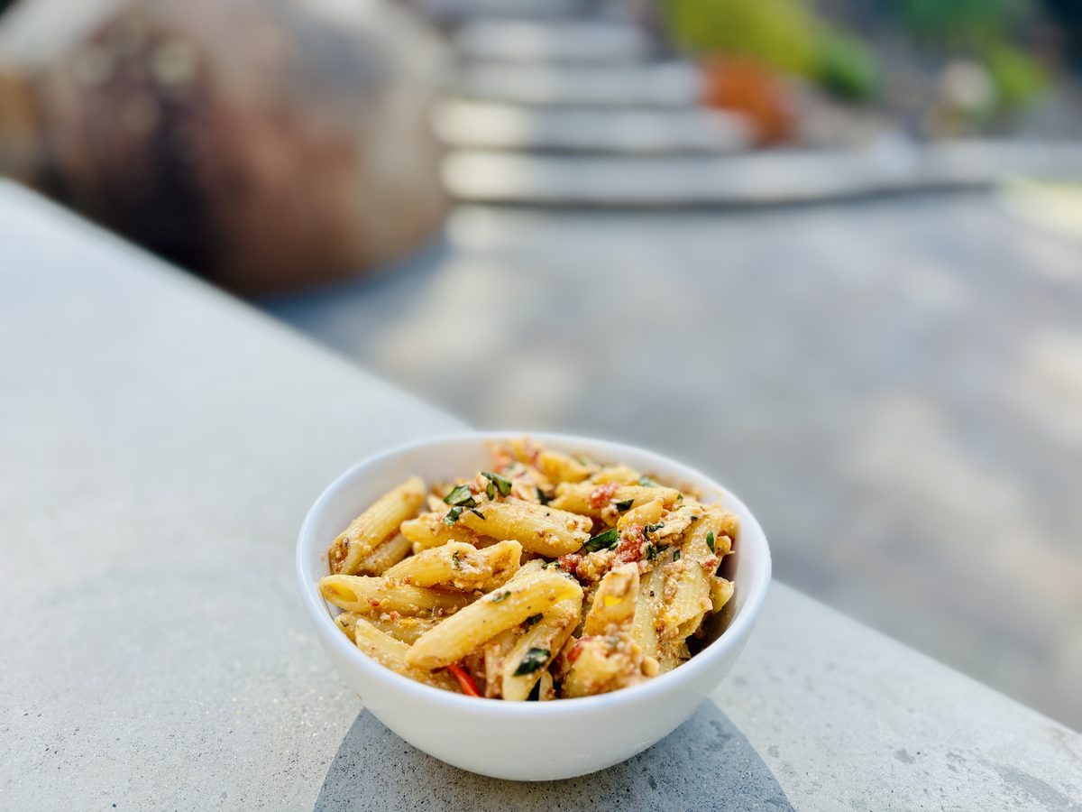 A small white bowl of penne tossed in a creamy sauce with herbs and bits of tomato, sitting on an outdoor stone ledge.