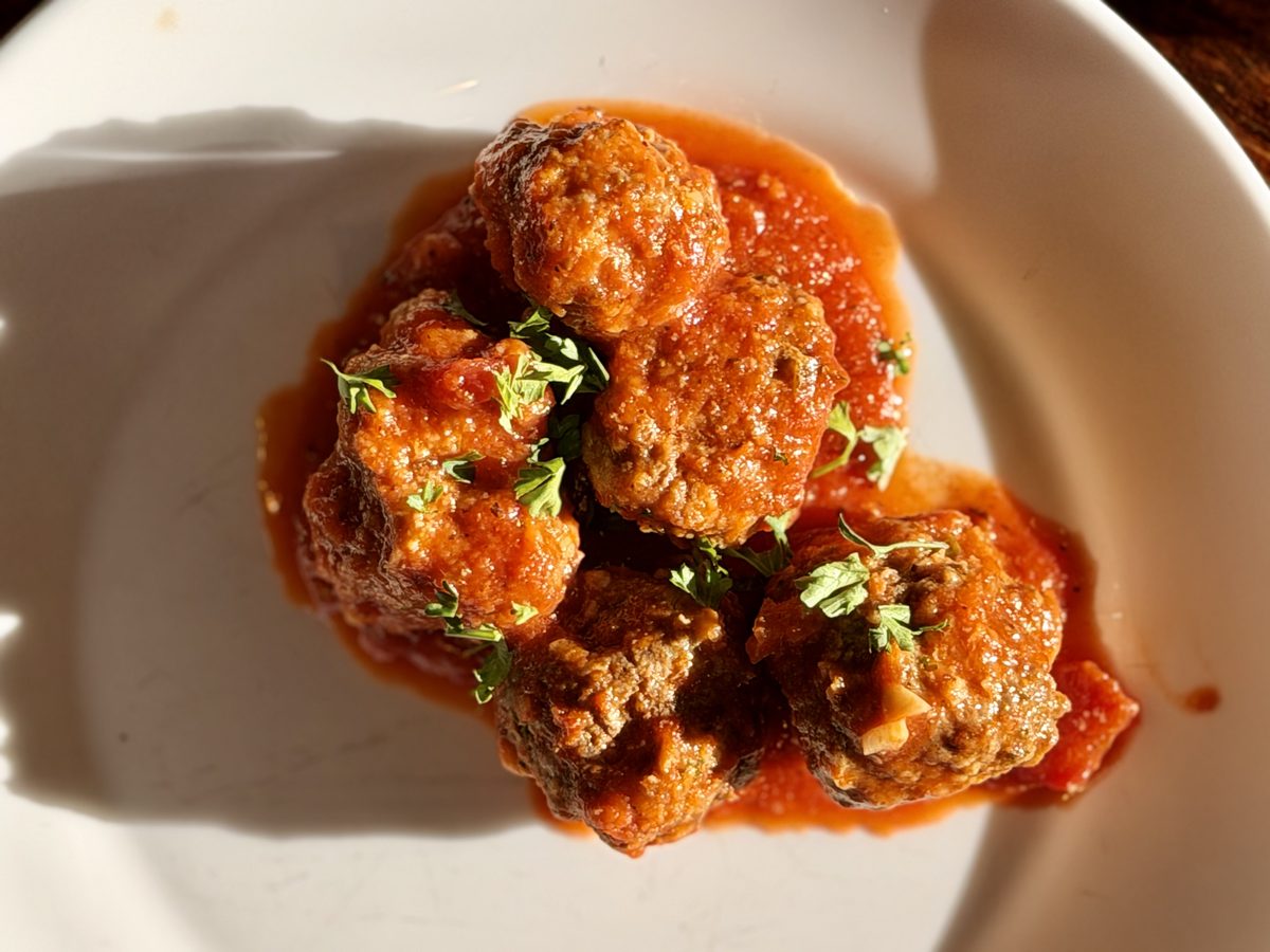 Close overhead view of four meatballs in tomato sauce on a white rimmed plate, garnished with torn basil, dramatic side light.