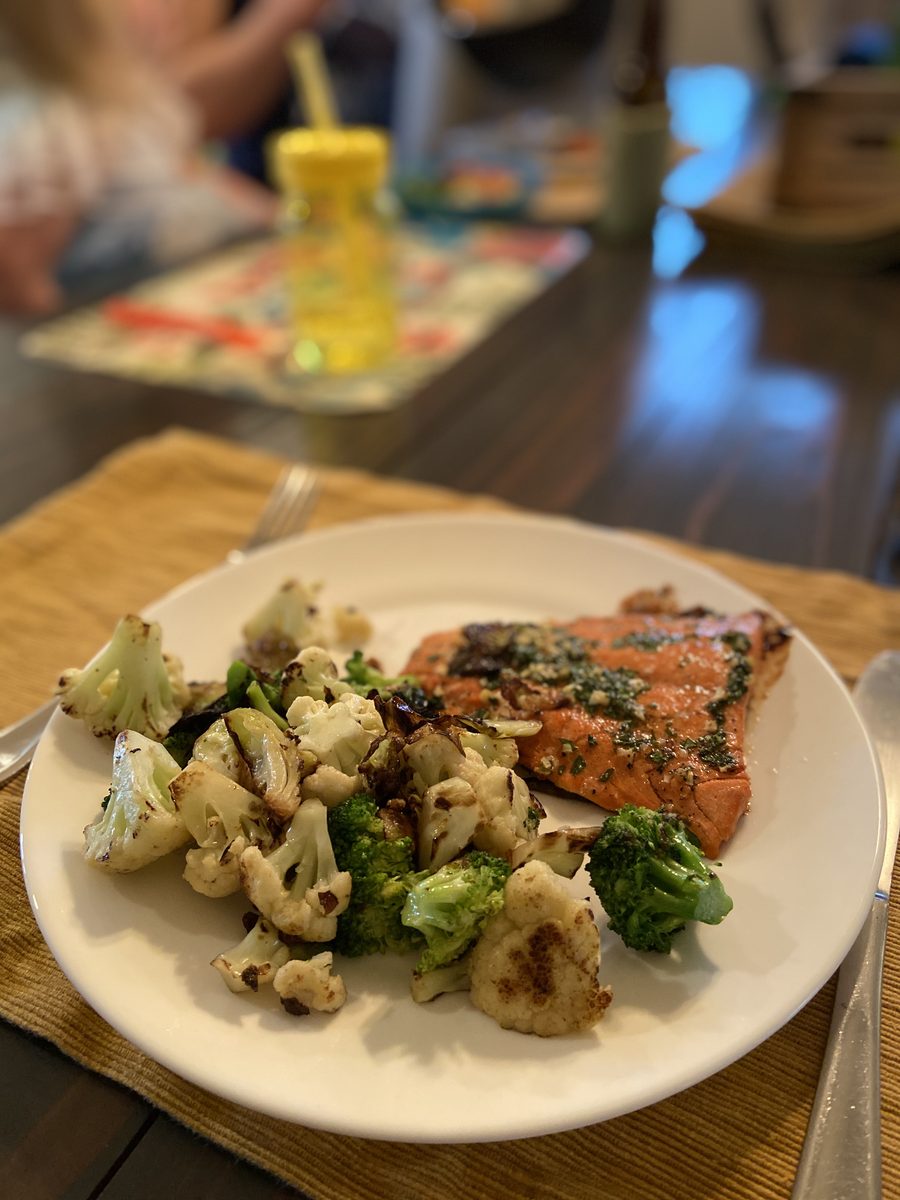A white plate holding a herb-topped baked salmon fillet with roasted cauliflower and broccoli, sitting on a placemat at a dim dini