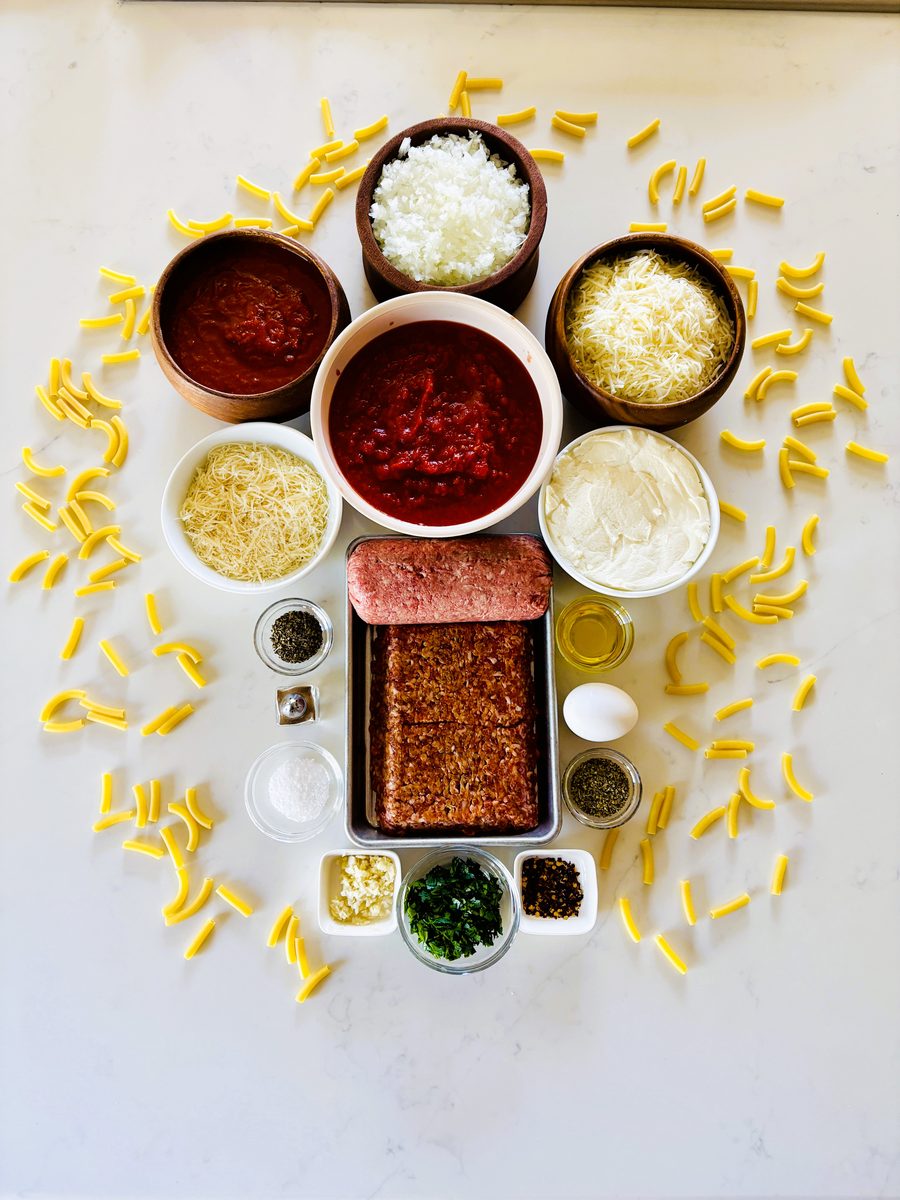 Overhead flat lay of baked-ziti ingredients on white marble: dried short pasta scattered in a ring around bowls of marinara, ricot