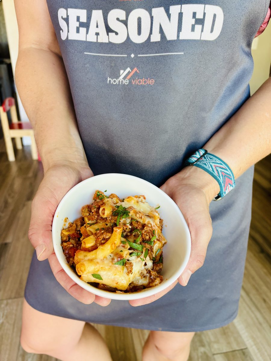 A person in a gray branded apron reading SEASONED holds a white bowl of baked ziti with ground meat sauce, melted cheese and herbs