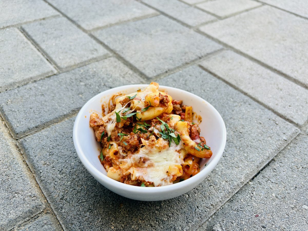 A white bowl of baked pasta in red sauce with melted cheese and herbs on top, set on paving stones; browned crumbles in the sauce 