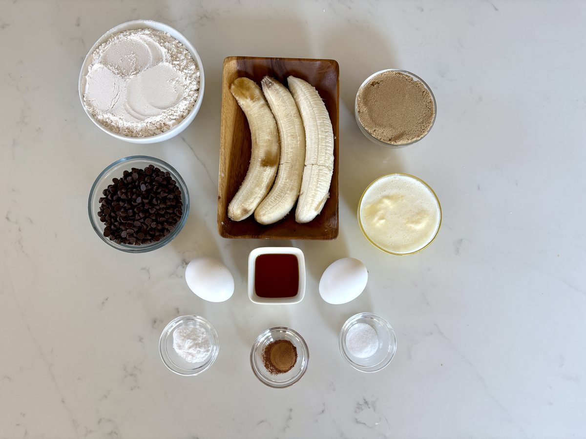 Overhead flat lay of banana-bread ingredients on bright white marble: peeled bananas on a wood board, bowls of flour, chocolate ch