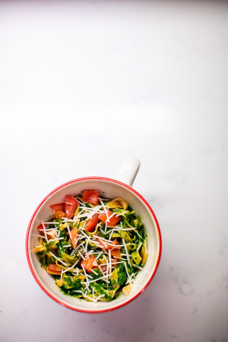 An overhead shot of a small red-rimmed mug bowl of green pesto pasta with wilted greens, halved cherry tomatoes and shredded chees