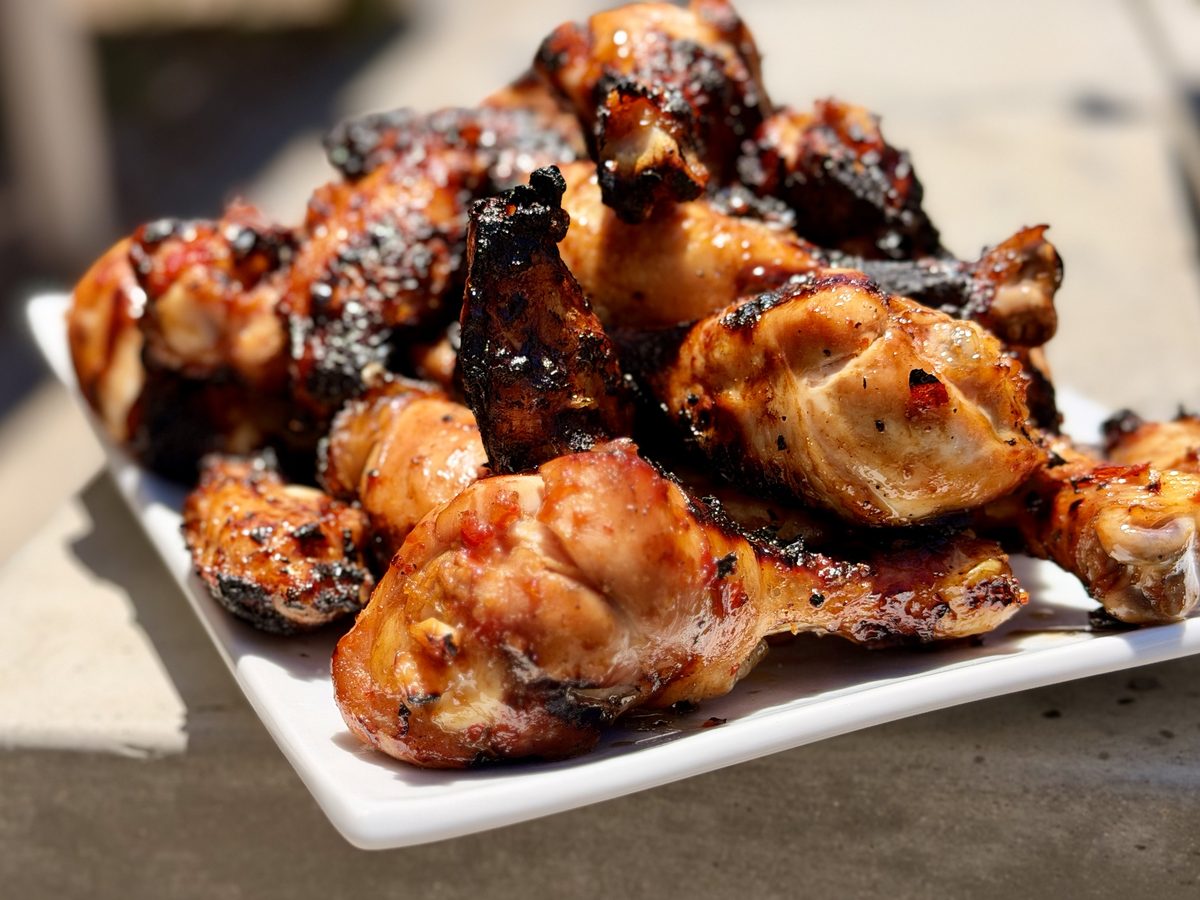 Close-up of a heaped white platter of glazed, char-marked barbecue chicken drumettes outdoors, sharp and sunlit.