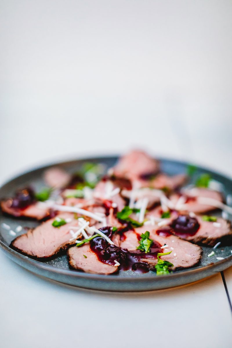 Sliced roast pork tenderloin fanned on a dark grey ceramic plate with dark cherry-berry sauce, shredded white cheese and fresh her
