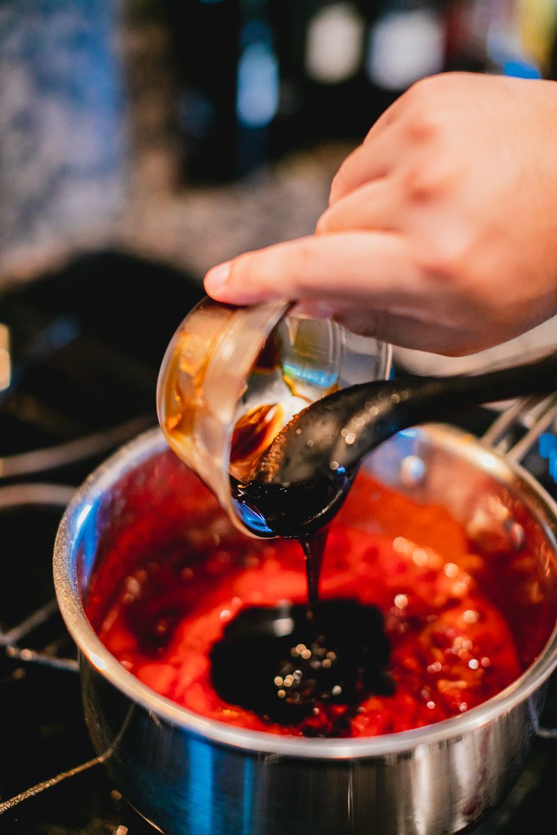A hand tips a small glass cup of dark molasses over a spoon into a saucepan of red sauce on the stove; blurred kitchen counter beh