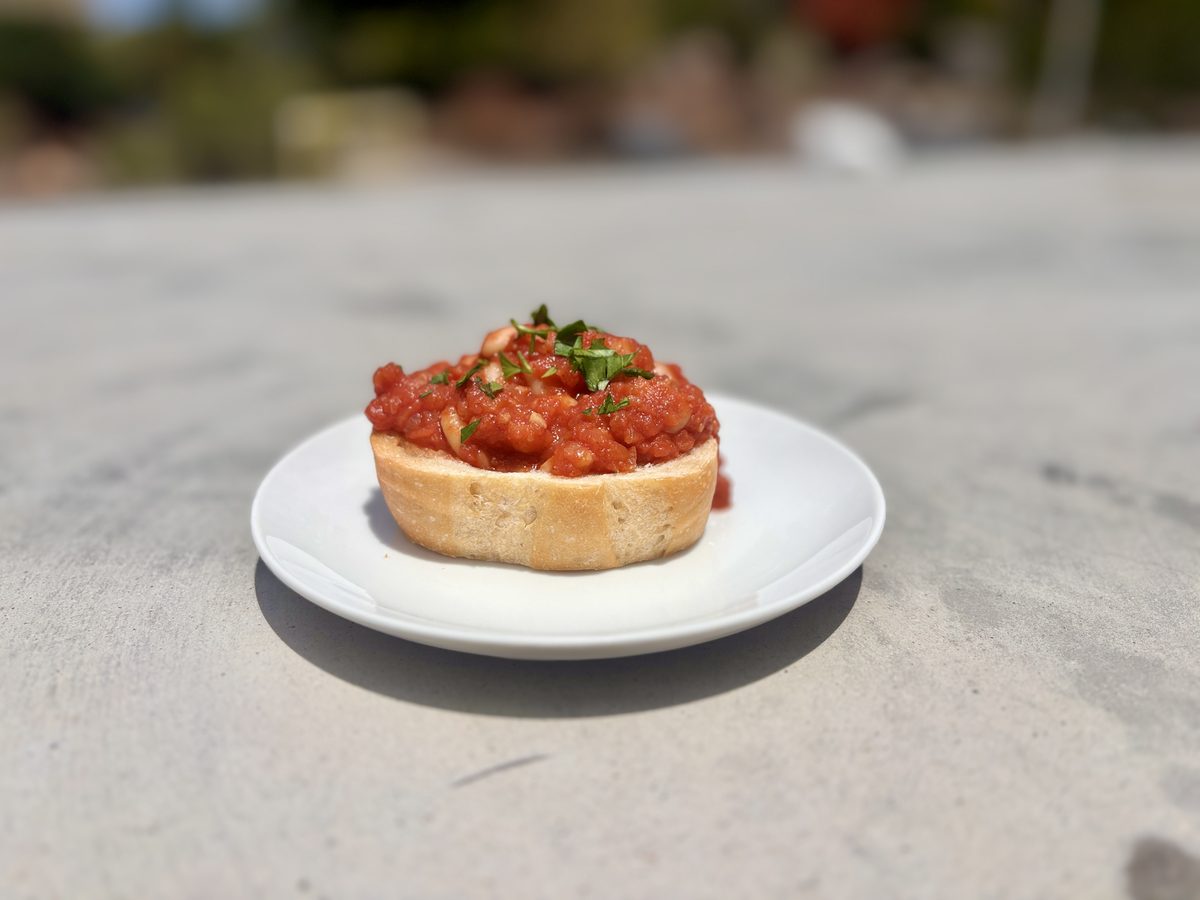 A single thick toast round piled with chunky red tomato-and-bean mixture and chopped herbs on a small white plate outdoors.
