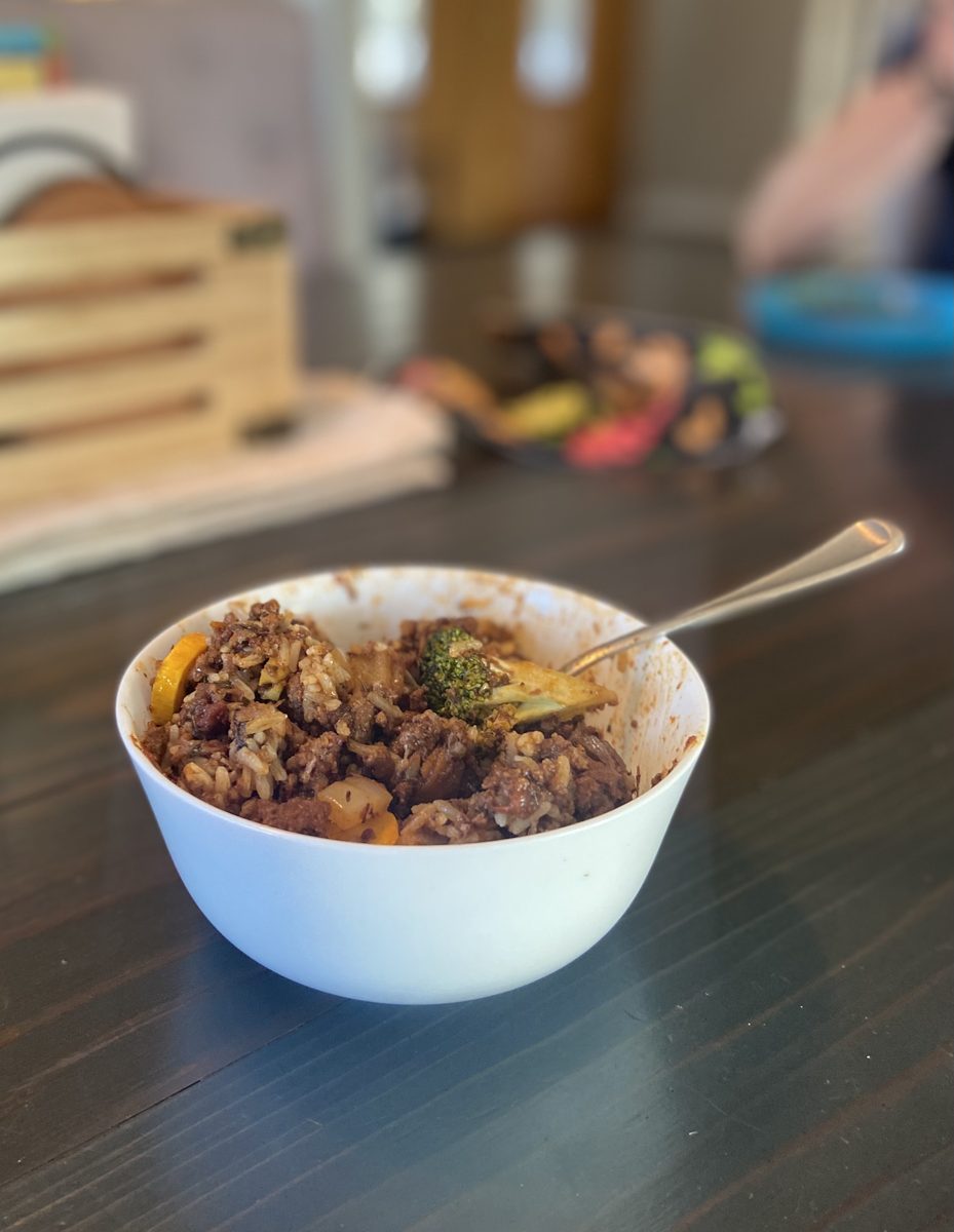 A white bowl of ground beef with rice and bits of vegetable, spoon resting in it, on a dark wood counter with a blurred kitchen an