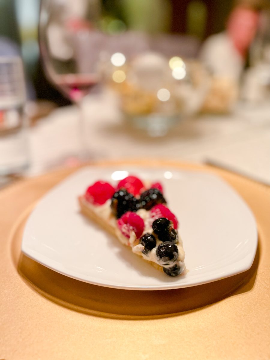 A slice of cream-filled tart topped with strawberries and blueberries on a white plate in a warmly lit restaurant.
