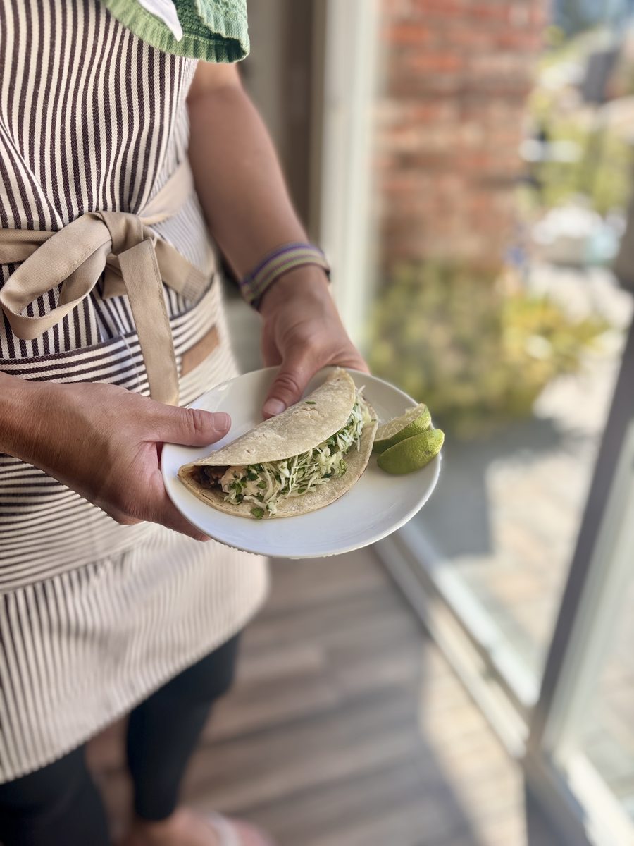 Hands hold a white plate with two folded black bean tacos filled with shredded cabbage and lime wedges, standing in a doorway.