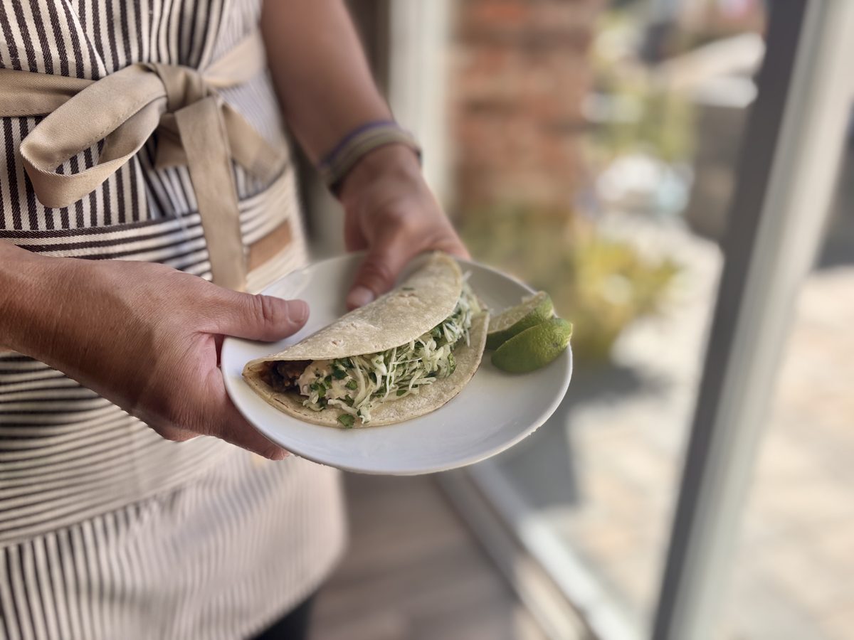 A person in a striped apron holds a plate of two assembled black bean and cabbage tacos with lime, backlit by a patio door.